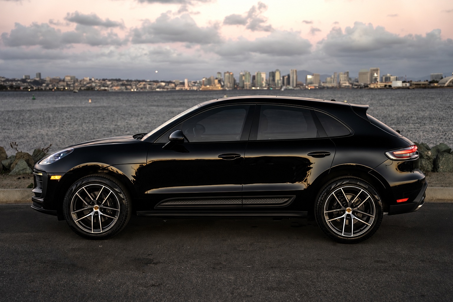 Side profile of the black Porsche Macan on silver multi-spoke wheels, San Diego skyline and water behind at dusk