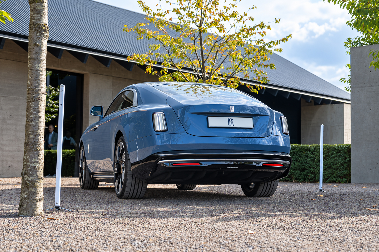 Rear three-quarter of the blue Rolls Royce Spectre showing the tapered tail and slim tail lamps, trees behind