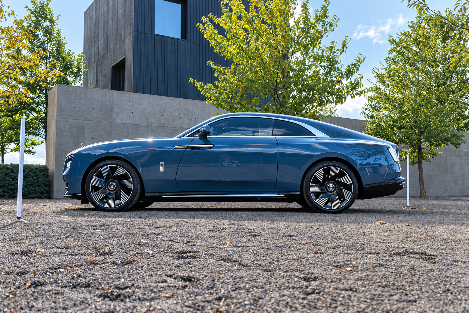 Side profile of the Salamanca Blue Rolls Royce Spectre on dark multi-spoke wheels, gravel courtyard and hedges behind