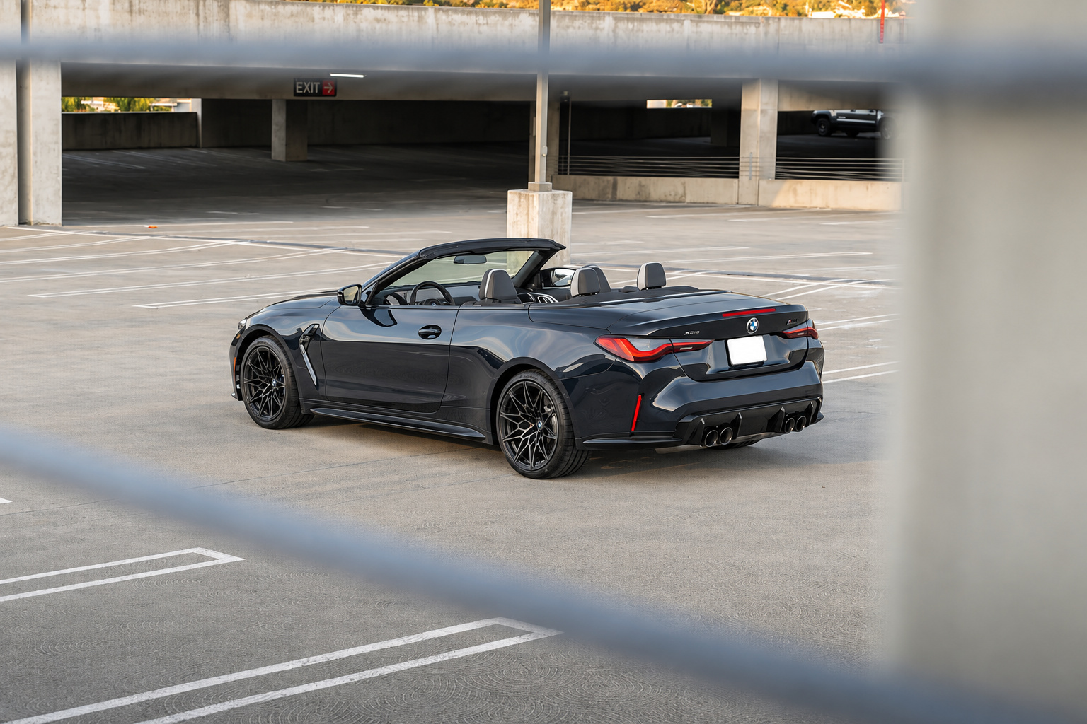 Rear three-quarter of the black M4 Competition Convertible, roof folded, quad pipes and black wheels on an open rooftop