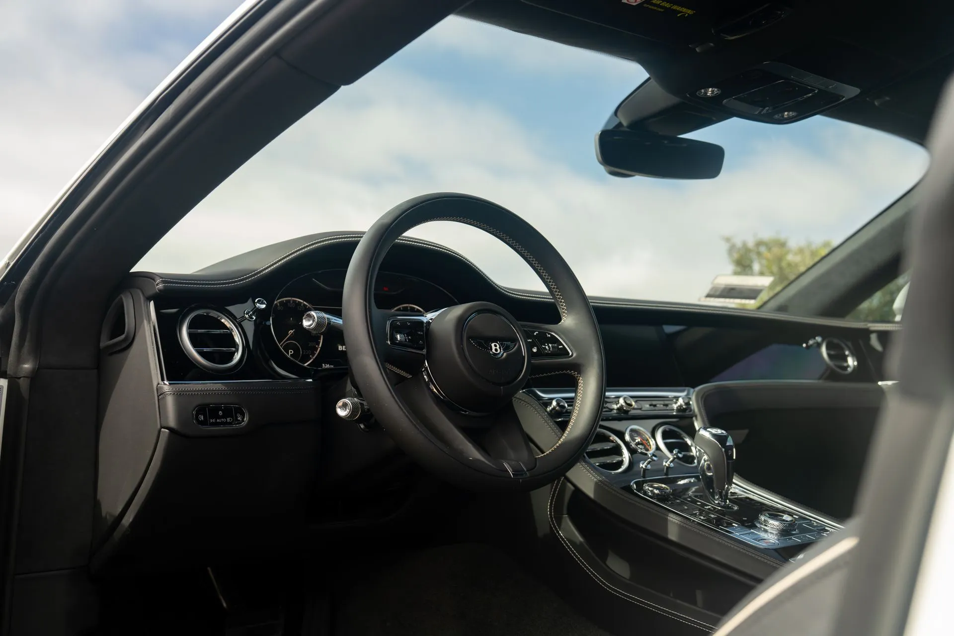 Driver's seat view of the white Continental GT, black leather steering wheel with the Bentley wings and the coast outside