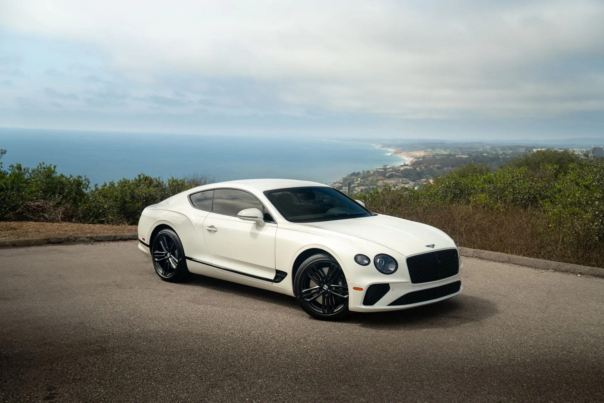 White Bentley Continental GT coupe with black wheels parked on a coastal overlook above the Pacific under an overcast sky