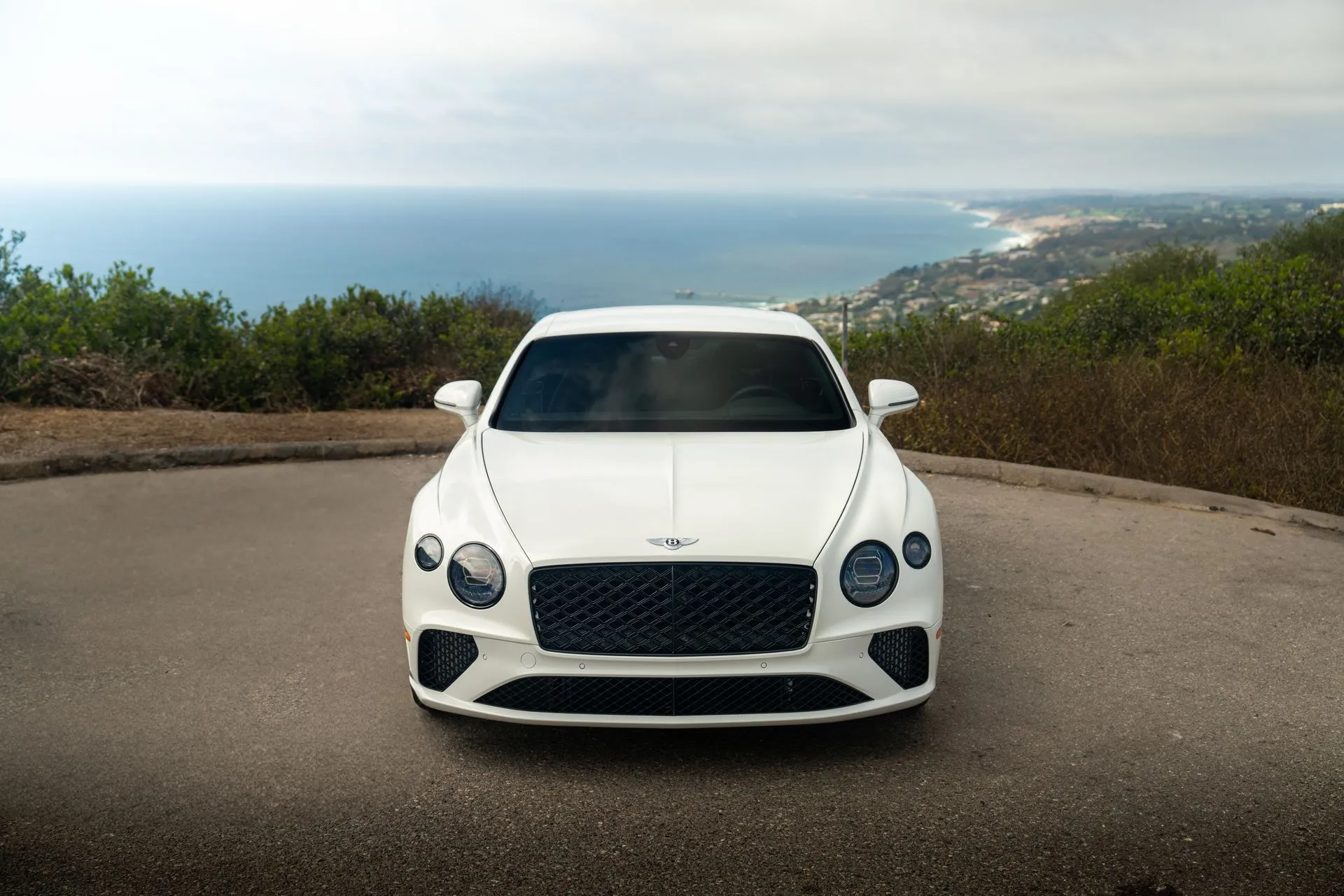 Head-on view of the white Bentley Continental GT, chrome grille and 4 round headlamps, with the coastline falling away behind