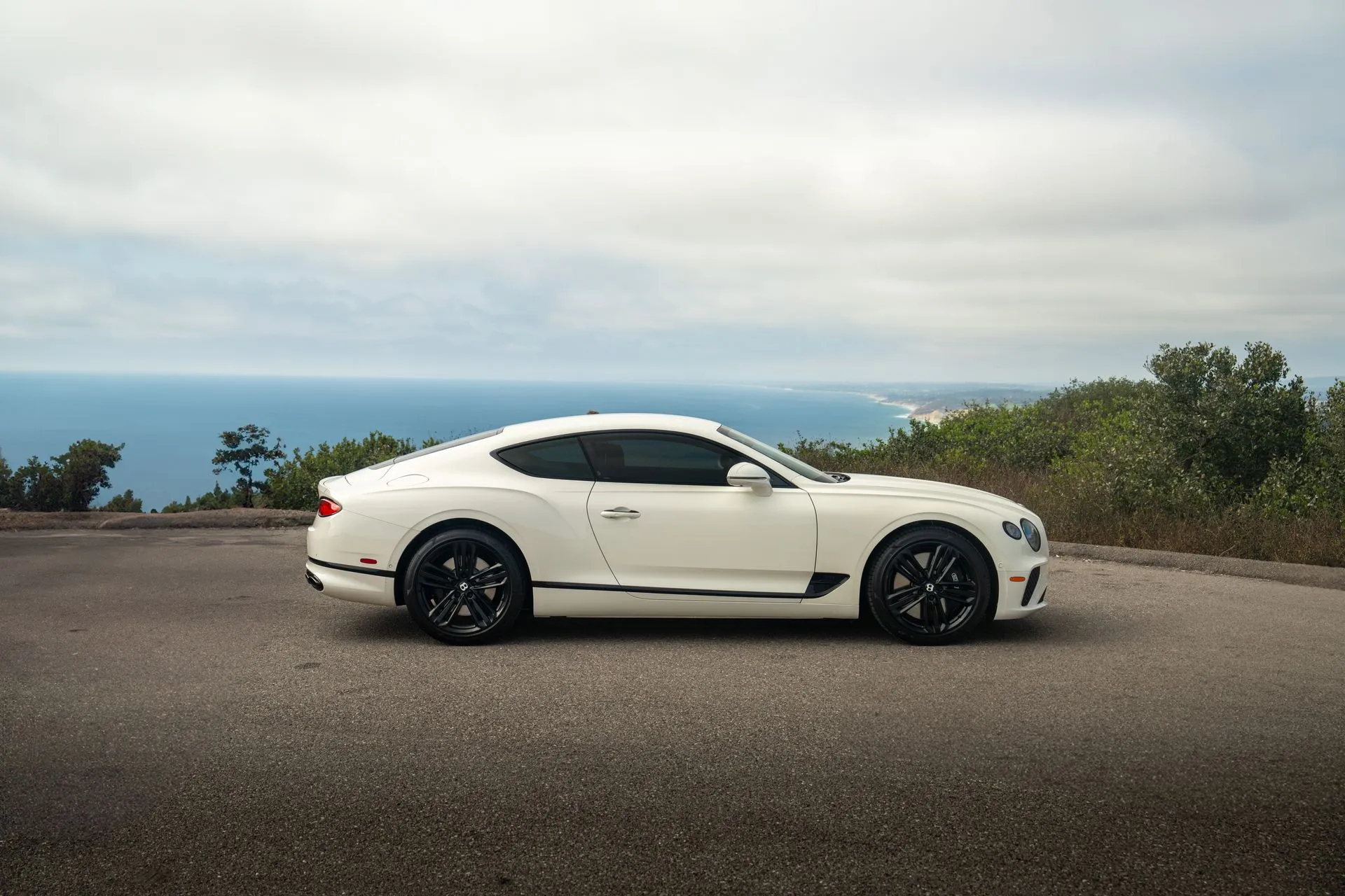 Side profile of the white Bentley Continental GT with gloss black wheels, the Pacific and a grey sky behind it