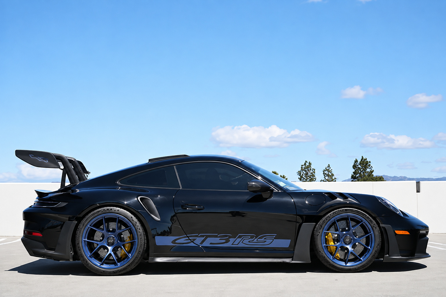 Side profile of the black Porsche 911 GT3 RS Weissach with the carbon wing and blue wheels, mountains behind