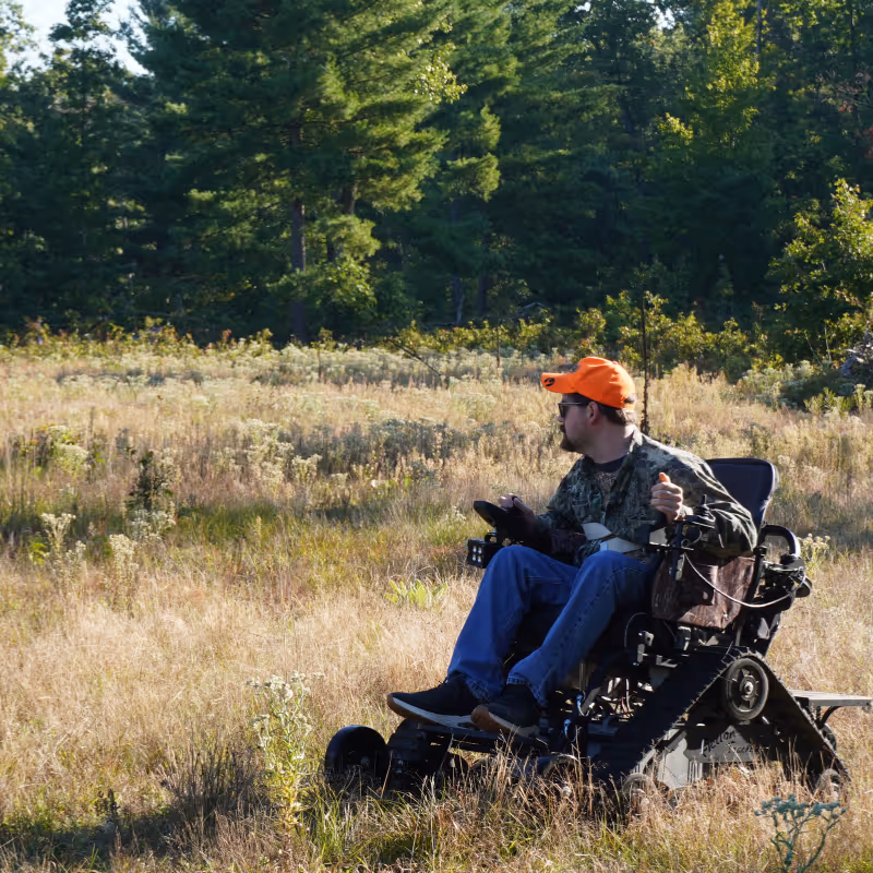Man in camouflage and orange cap sitting in off-road wheelchair in grassy field near trees.