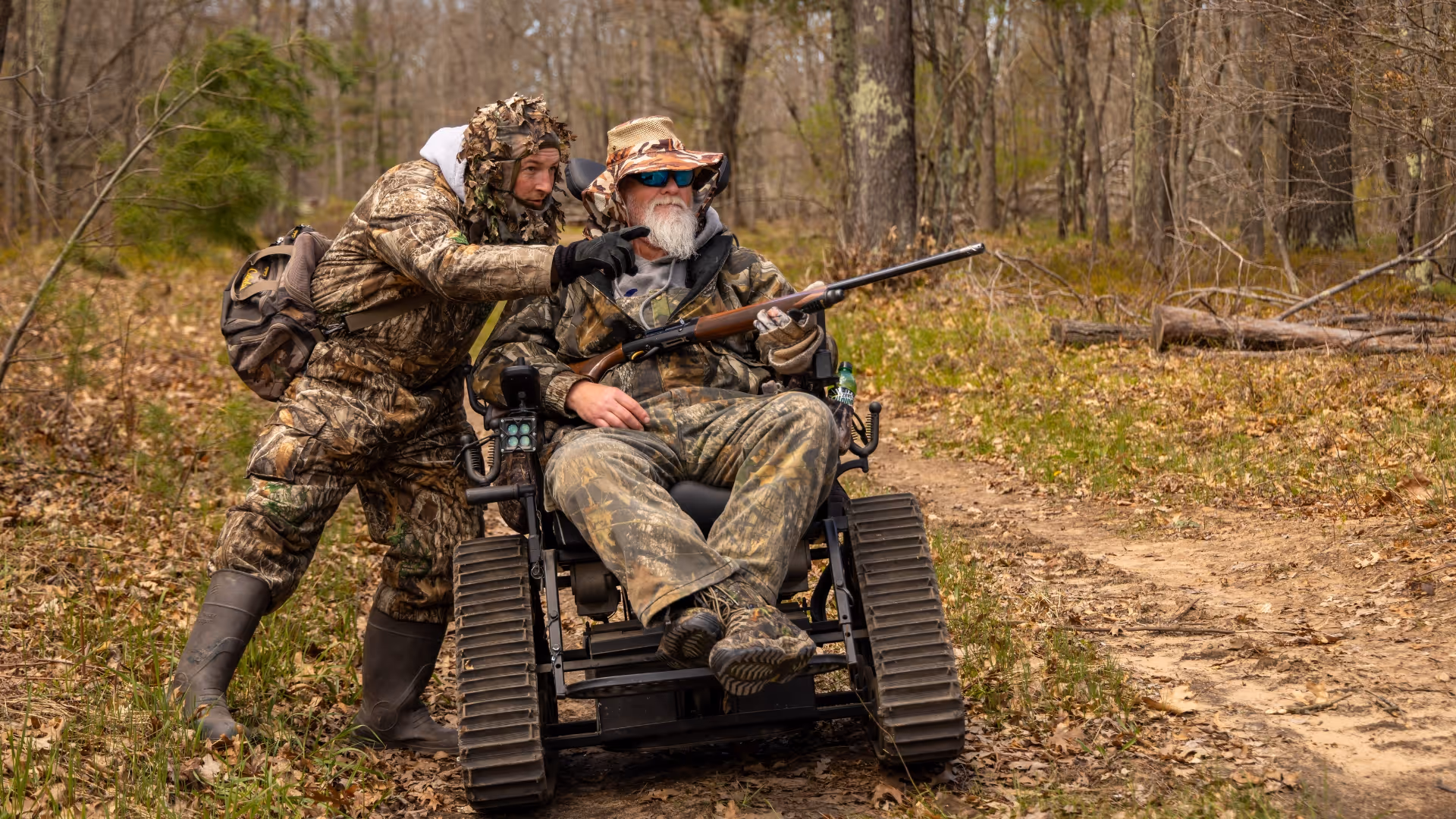 Two hunters in camouflage, one in a tracked wheelchair holding a rifle, the other pointing ahead in a forest.