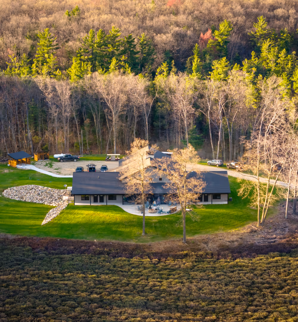 A large lodge surrounded by green lawn and forest with several parked cars and outdoor seating area.