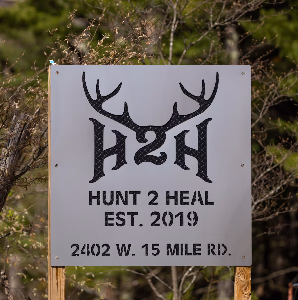 Metal sign with antlers logo and text: Hunt 2 Heal Est. 2019, 2402 W. 15 Mile Rd.