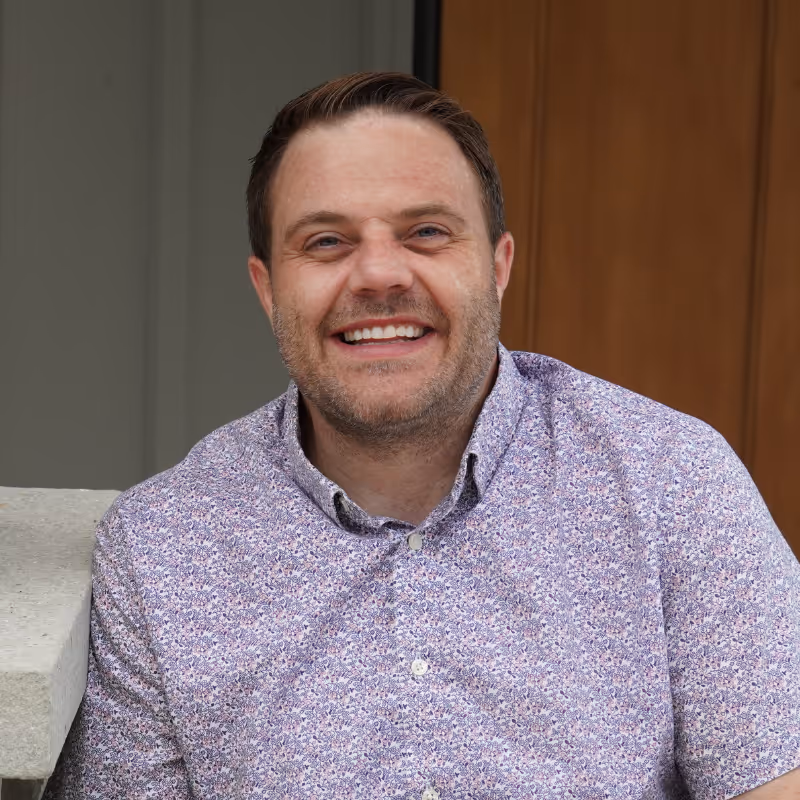 Smiling man wearing a floral button-up shirt leaning on a concrete ledge indoors.
