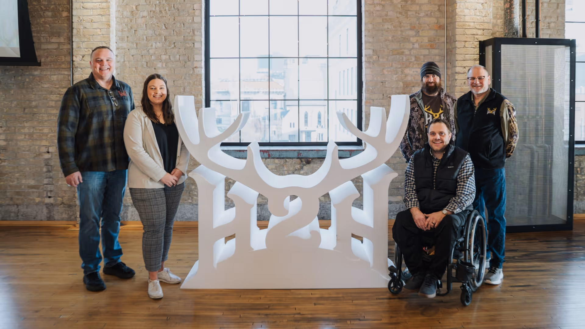 Five people posing around large white H2H sign with antlers in a room with brick walls and large windows.