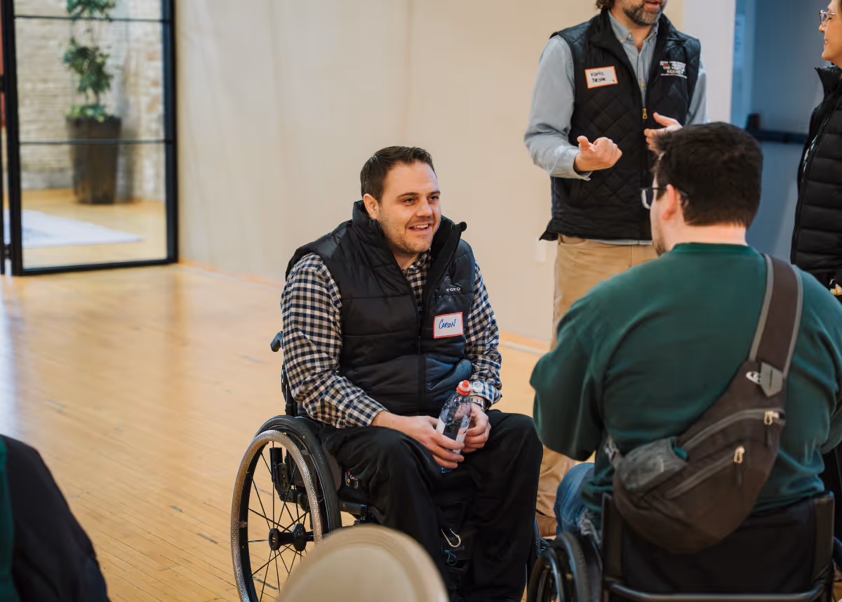 Two men in wheelchairs talking and smiling indoors with two standing people in conversation nearby.