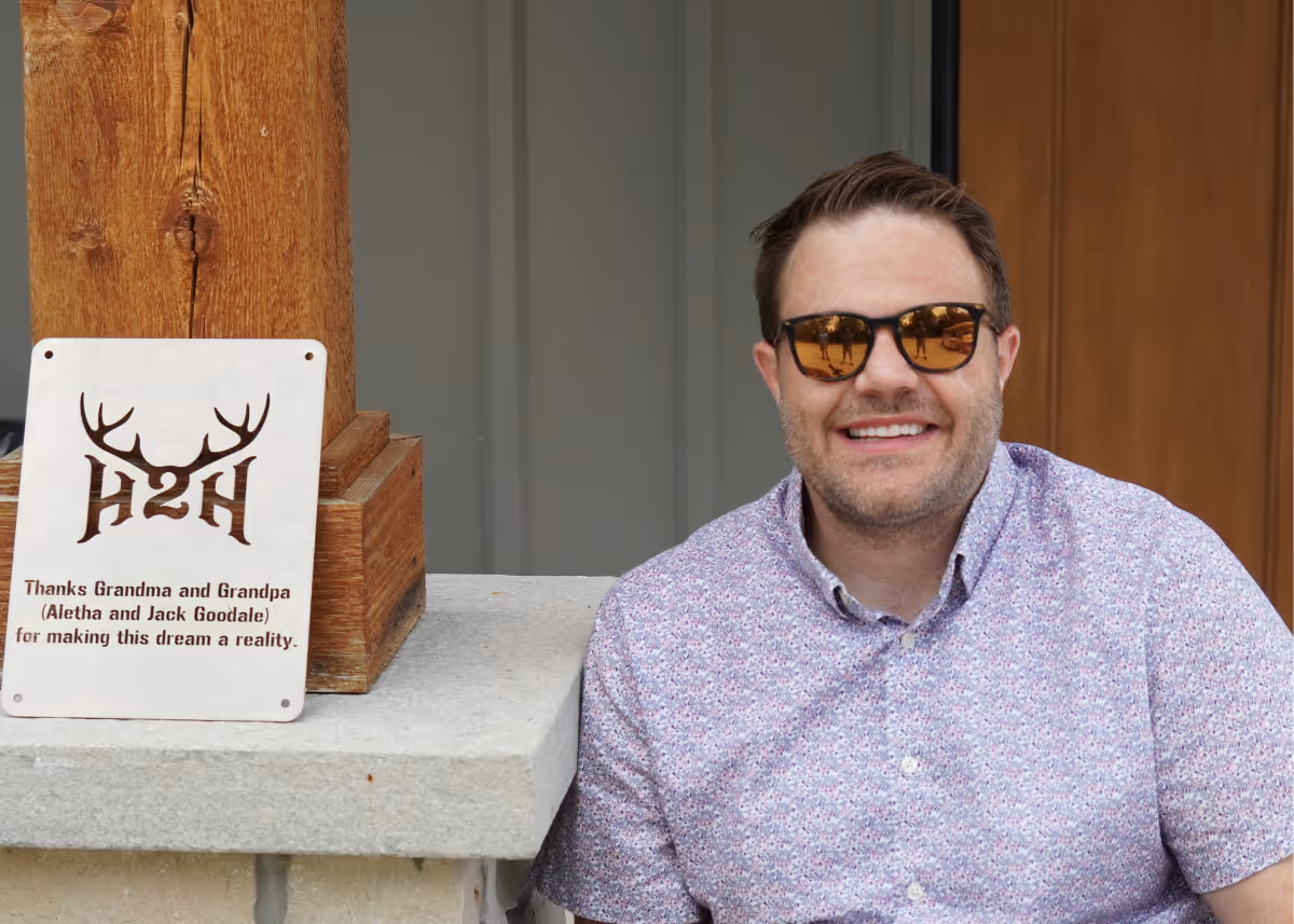 Smiling man wearing sunglasses and a patterned shirt sitting next to a wooden post with a thank you plaque.