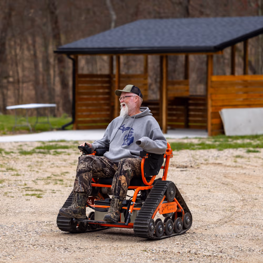 Man with a white beard in camo pants uses an orange tracked wheelchair outdoors near a wooden shelter.