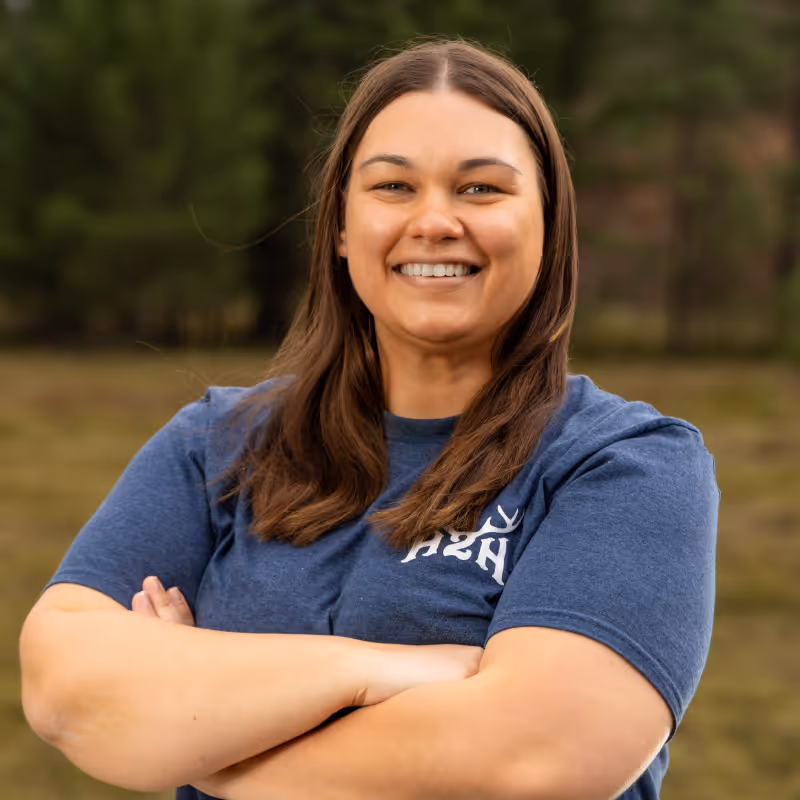 Smiling woman with long brown hair wearing a blue shirt with white lettering outside with arms crossed.
