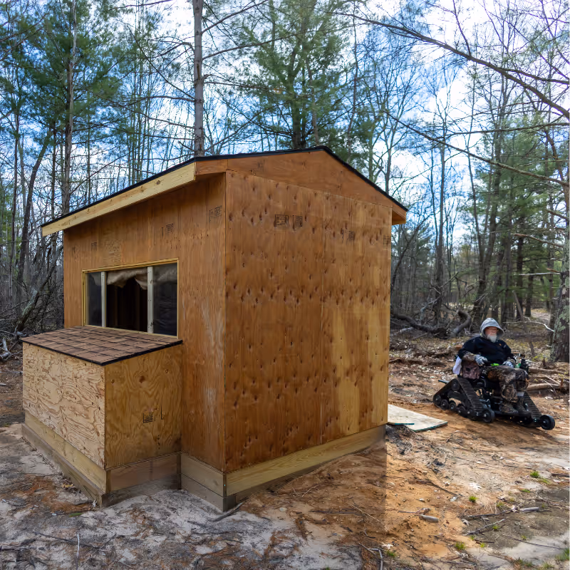 Person in mobility chair with tracks by unfinished plywood shed in forest clearing.
