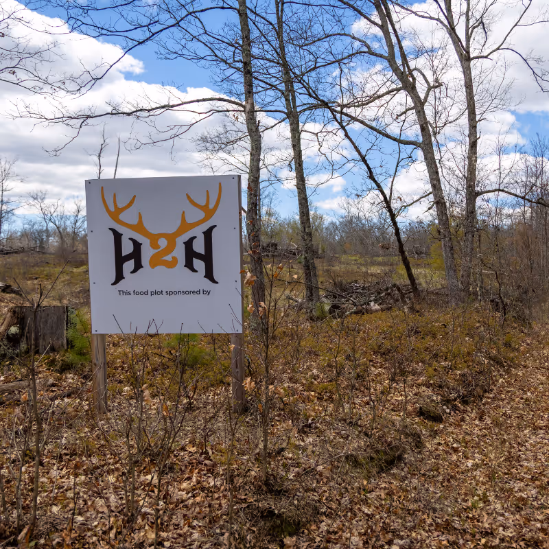 Sign with antlers and H2H logo in a leaf-covered clearing with bare trees under a partly cloudy sky.
