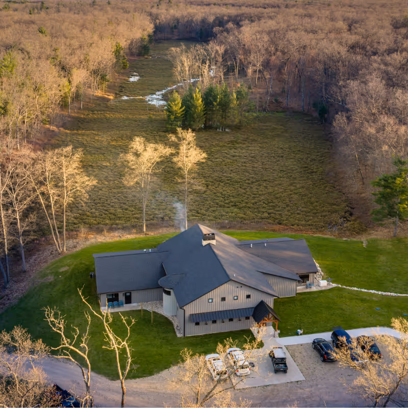 A large lodge with a dark roof surrounded by green lawn and forest in late autumn or early winter.
