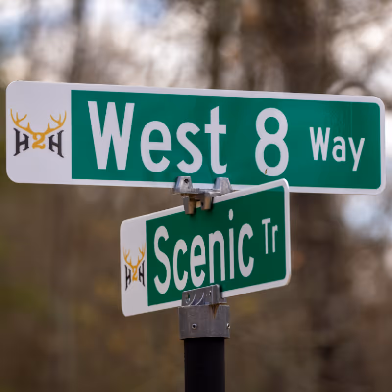 Street signs reading West 8 Way and Scenic Tr on a black pole with a blurred natural background.