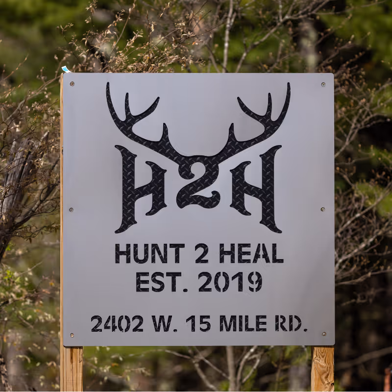 Sign with deer antlers logo reading 'H2H Hunt 2 Heal Est. 2019 2402 W. 15 Mile Rd.' outdoors.