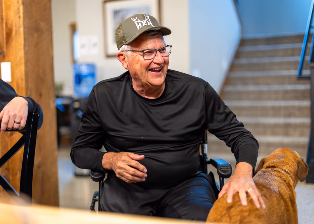 Smiling elderly man in black shirt and cap sitting in a wheelchair petting a brown dog indoors.