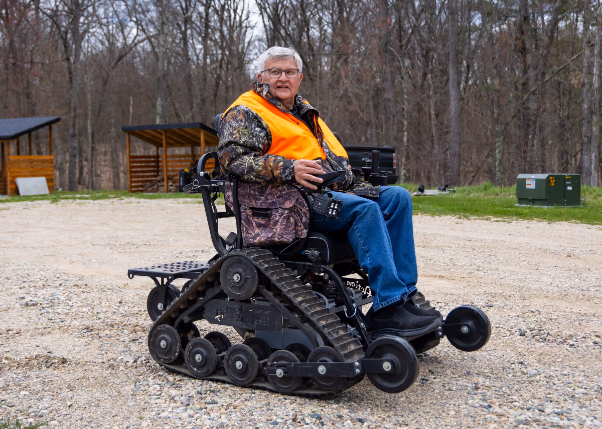 Man in camouflage jacket and orange vest sitting in a tracked all-terrain wheelchair outdoors.