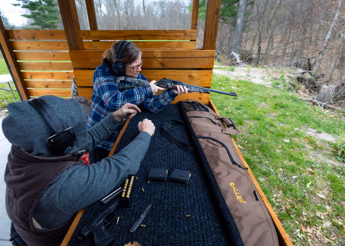 Woman aiming a rifle while a man beside her instructs at an outdoor shooting range.