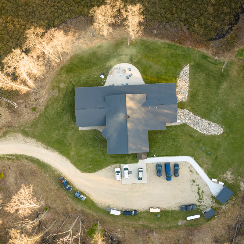 Aerial view of a T-shaped building surrounded by grass, trees, a curved driveway, and parked cars.