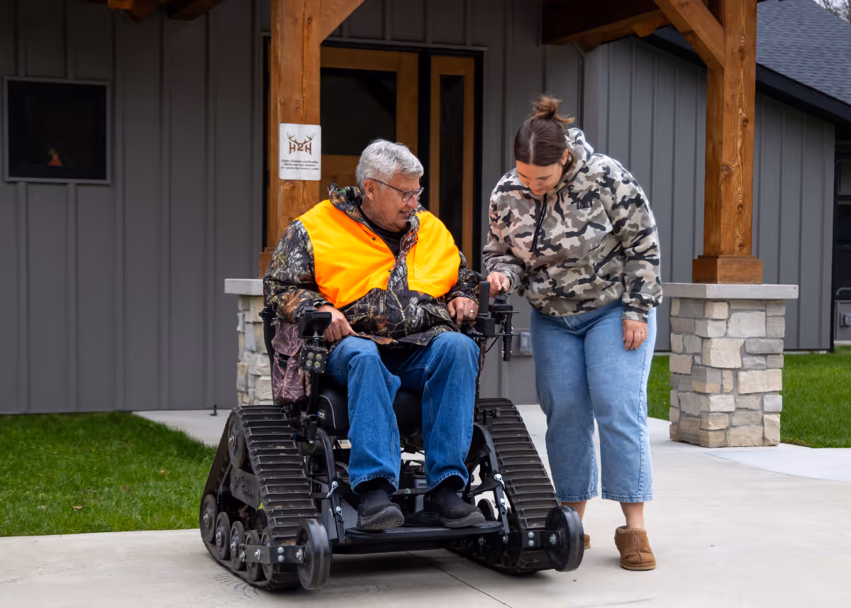 Older man in tracked wheelchair with bright orange vest, assisted by woman in camo hoodie outside house.