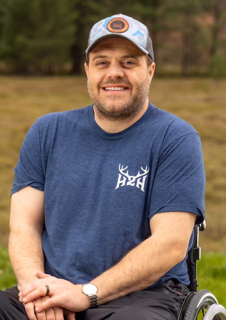 Smiling man in a wheelchair wearing a blue H2H T-shirt and camo cap outdoors.
