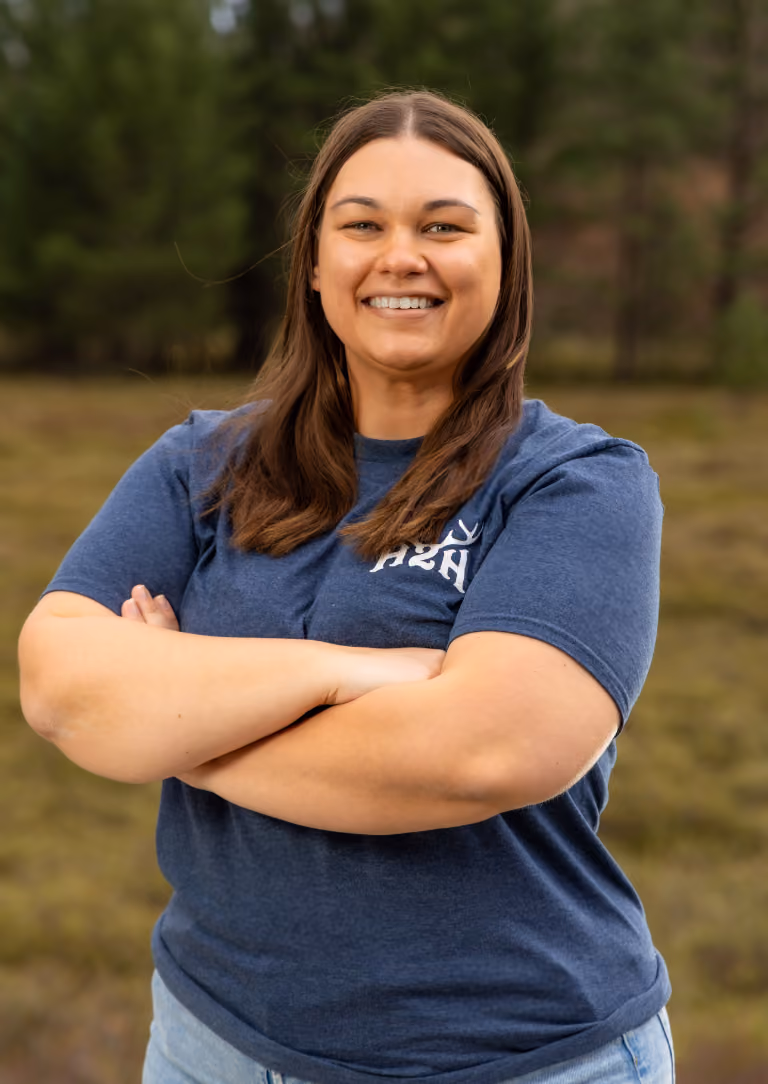 Smiling woman with straight brown hair in blue shirt and jeans standing outdoors with arms crossed.