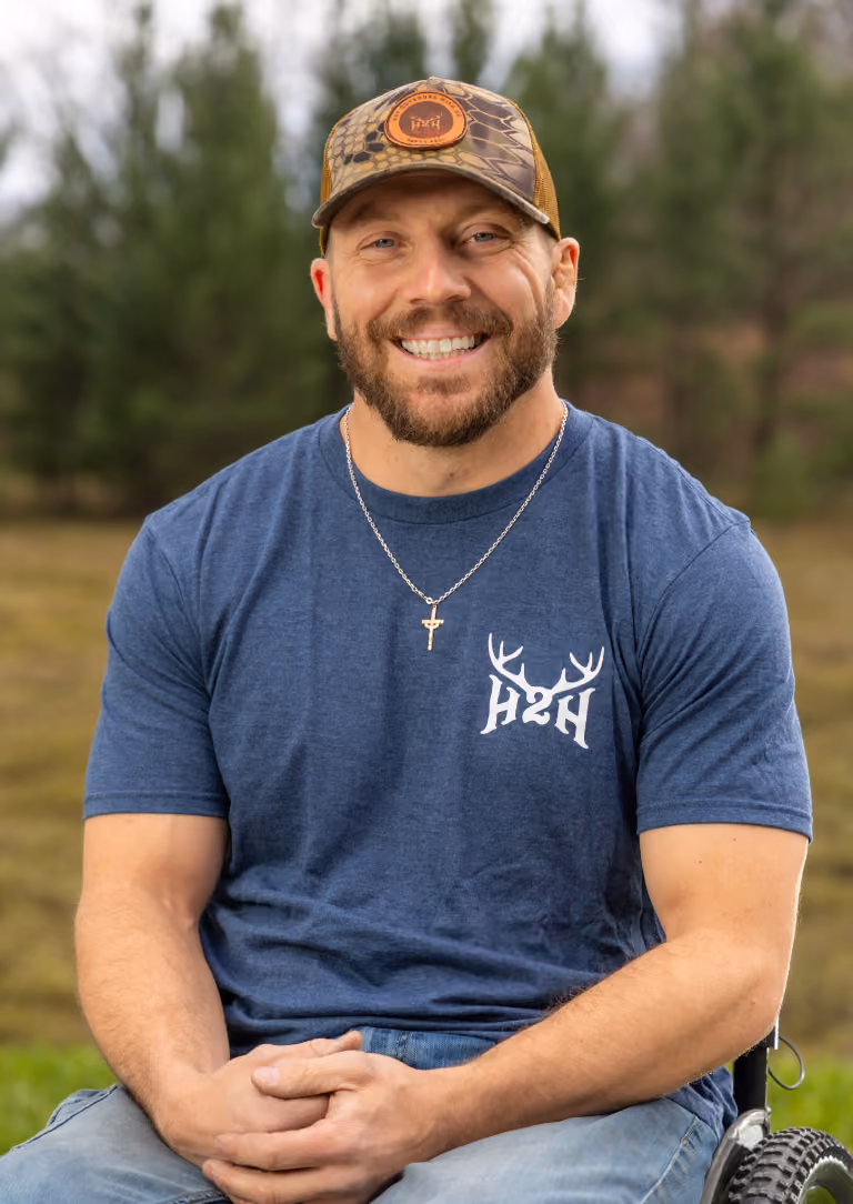 Smiling man with beard wearing a blue H2H shirt and a camouflage hat outdoors.
