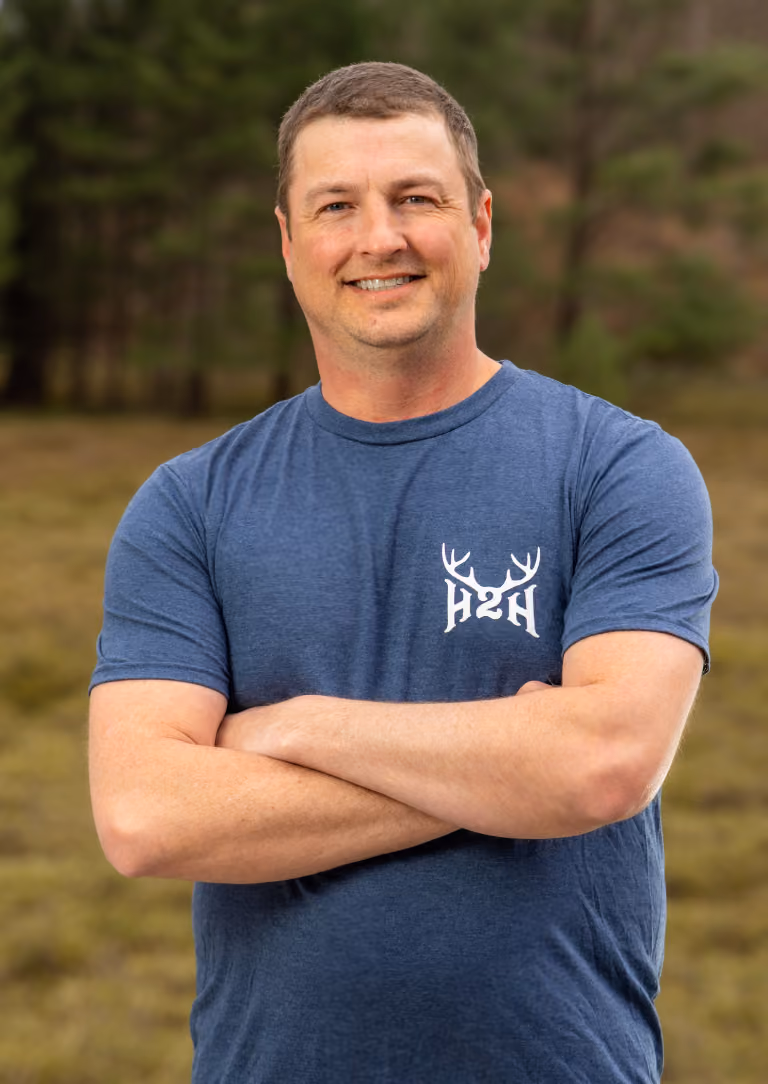 Smiling man with short hair in blue t-shirt with H2H logo, standing outdoors with arms crossed.