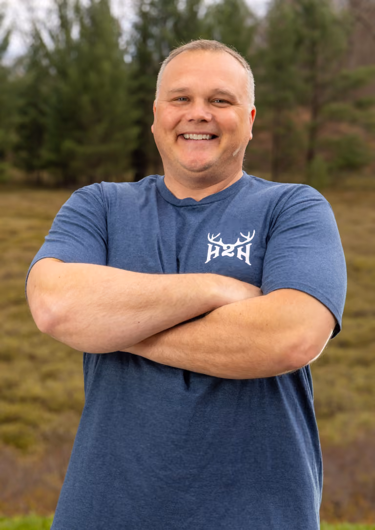 Smiling man in blue H2H t-shirt stands with arms crossed outdoors with trees in background.