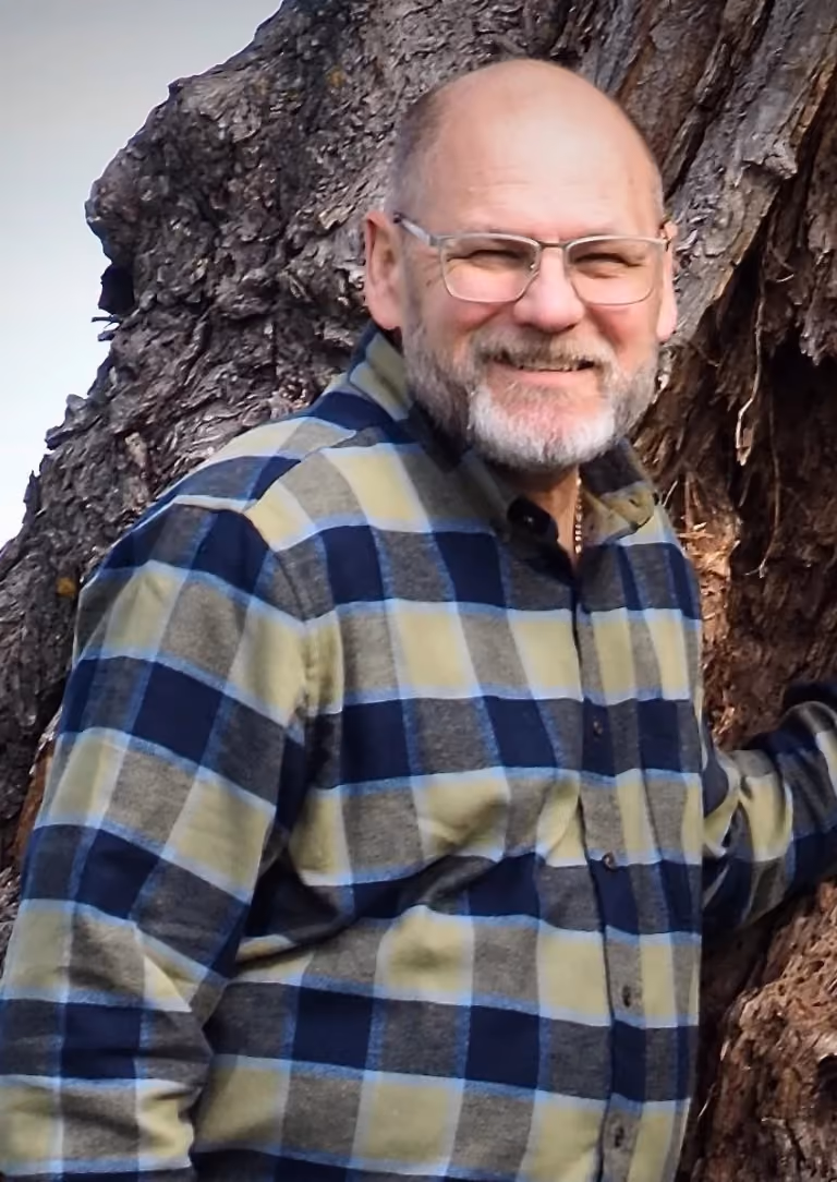 Smiling bald man with glasses wearing a plaid shirt standing by a large tree trunk.