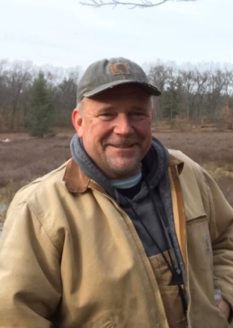 Man wearing a cap, tan jacket, and hoodie smiling outdoors in a wooded area.