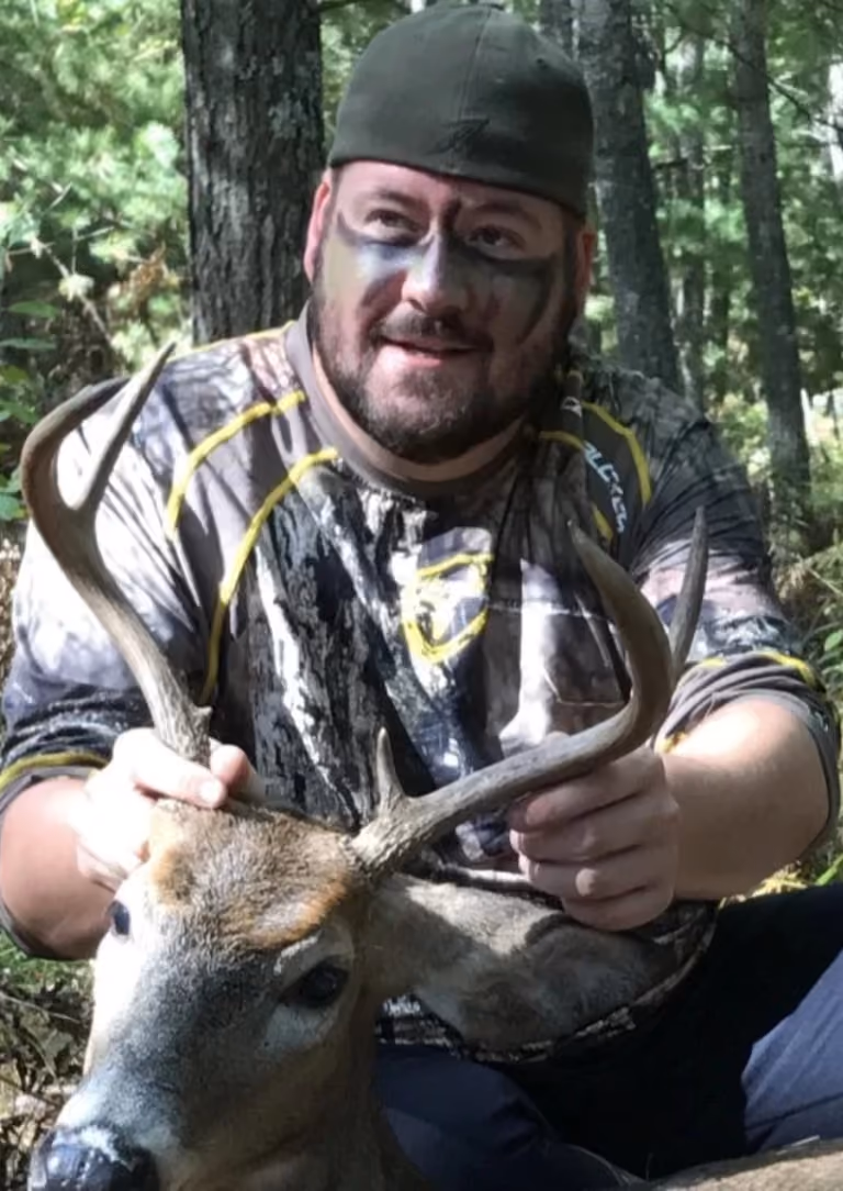 Man with face paint and camo clothing holding antlers of a deer in a wooded area.
