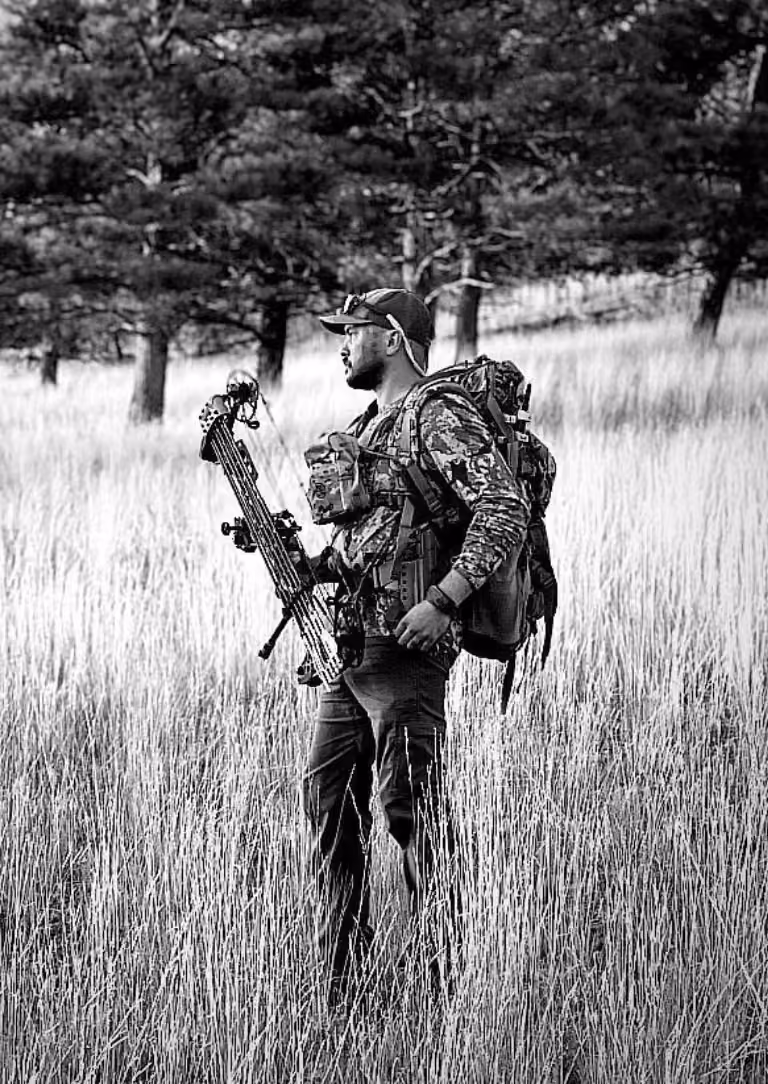 Man in camouflage gear holding a crossbow standing in tall grass near pine trees.
