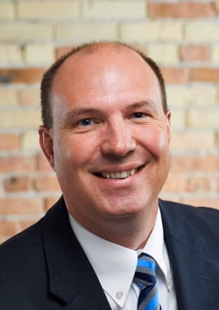 Smiling bald man in a dark suit with white shirt and blue striped tie against a brick wall.
