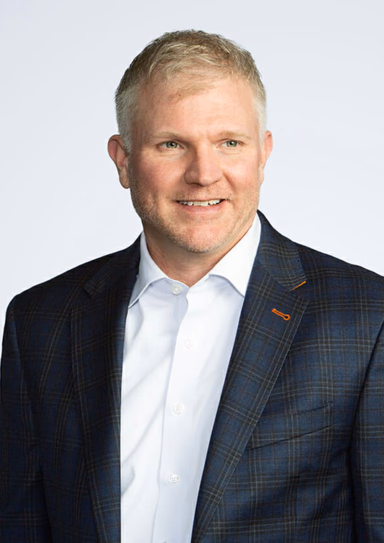 Smiling man with short blond hair wearing a blue plaid suit jacket and white shirt.