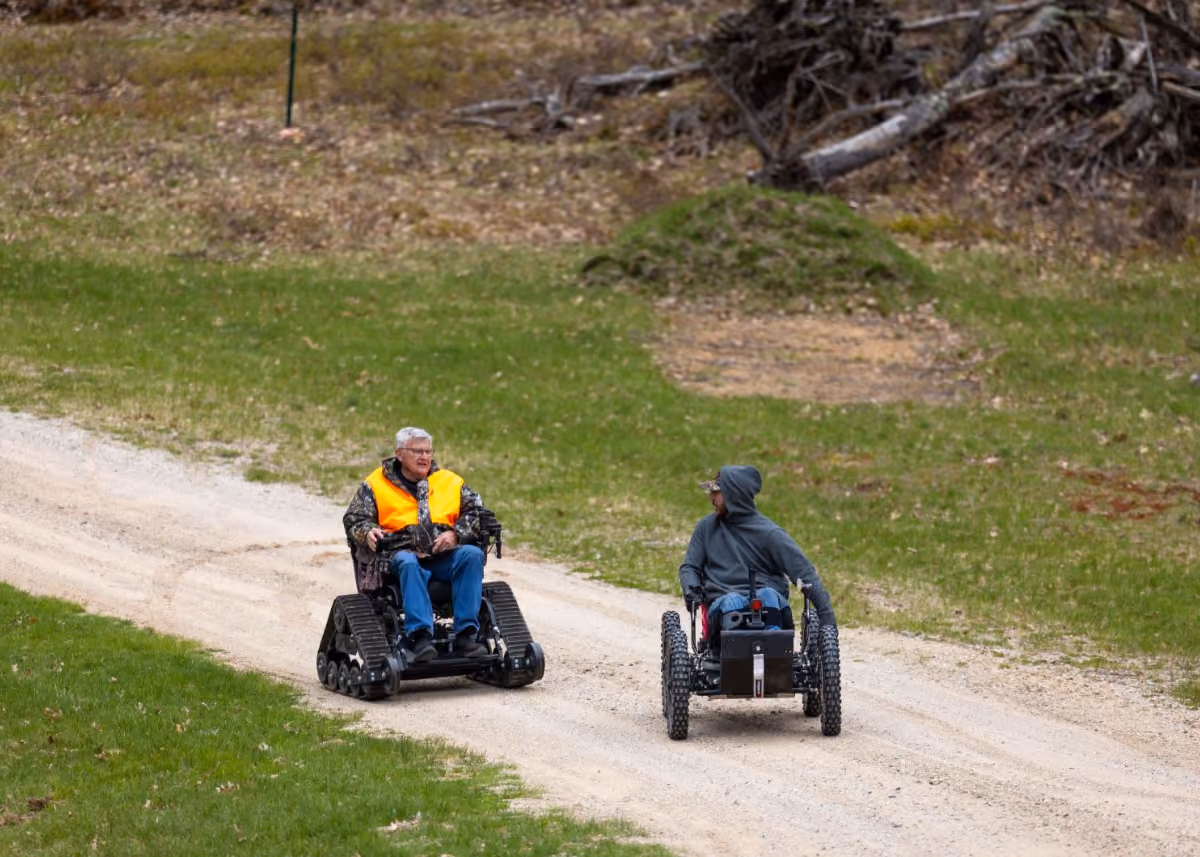 Two men on all-terrain wheelchairs on a dirt path surrounded by grass and trees.