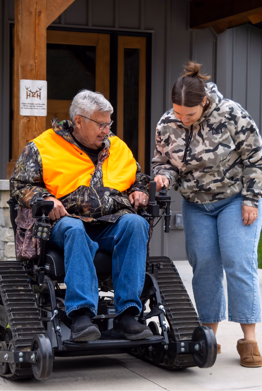 Older man in camouflage and orange vest on tracked wheelchair, woman adjusts controls outside building.