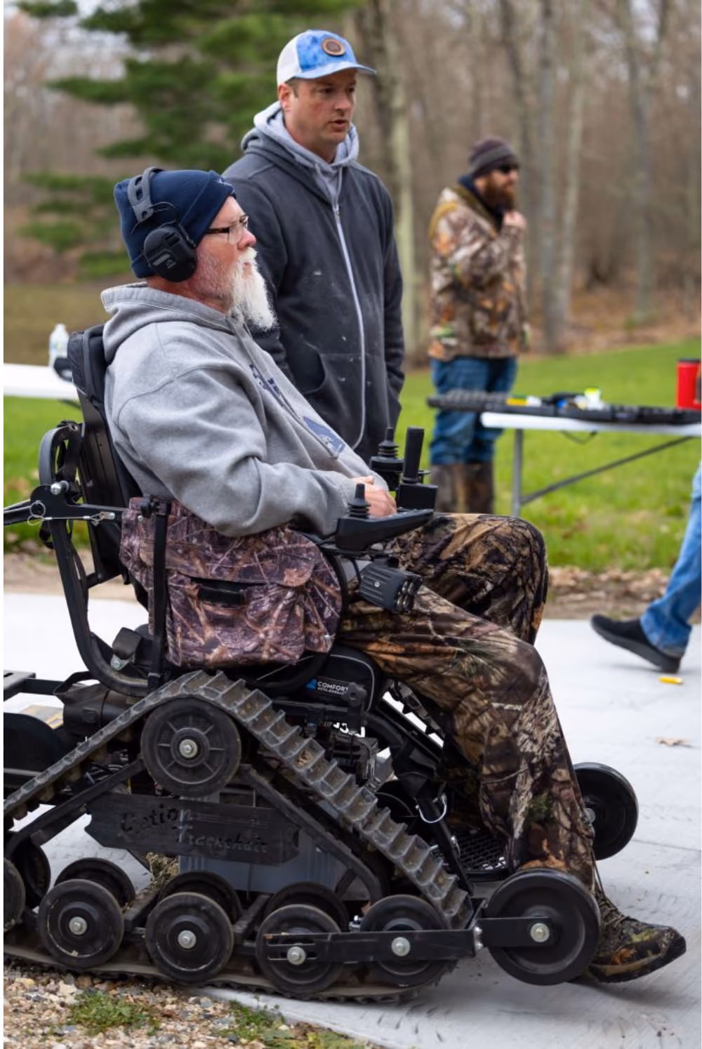 Man with white beard in camouflage pants using a tracked wheelchair outdoors with two other men nearby.