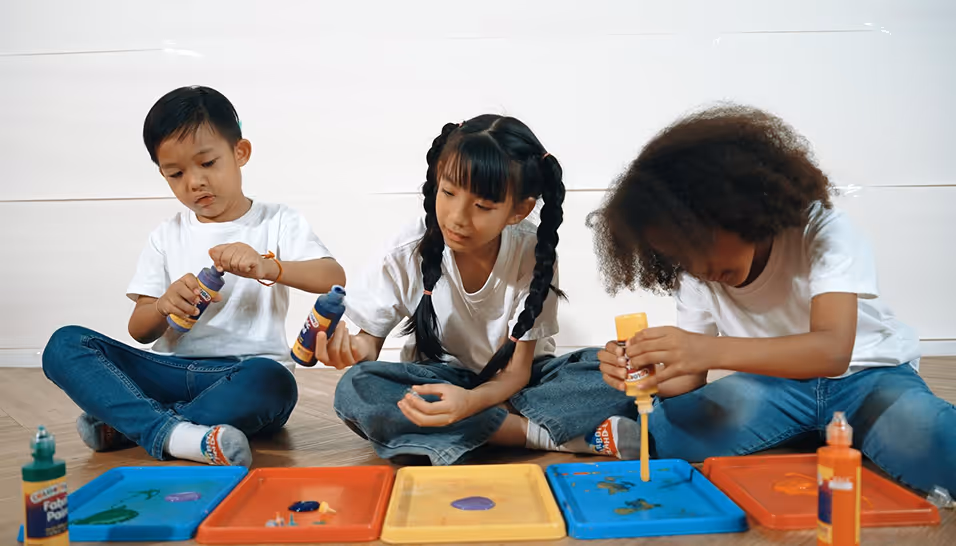 Three children sitting cross-legged painting on trays with colorful paint bottles indoors.