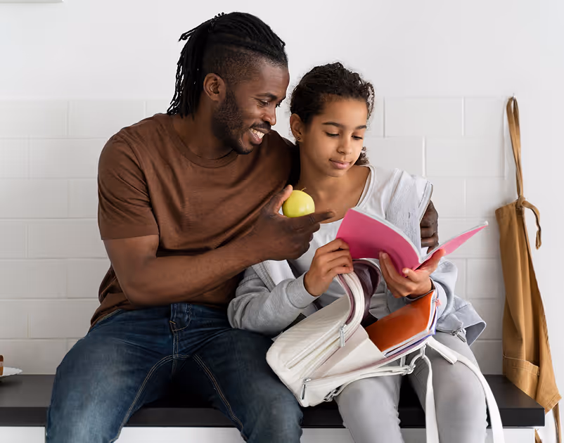Smiling man holding a green apple, sitting beside a girl who is reading a pink notebook from her open backpack.