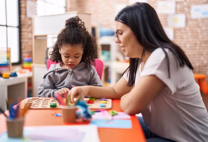 A woman with glasses helping a young girl with curly hair play a wooden educational puzzle at a colorful table in a bright classroom.