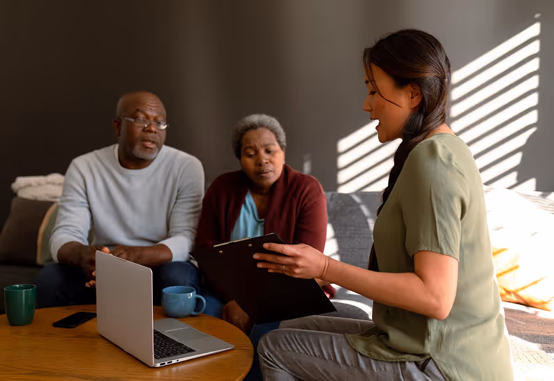 A woman holding a clipboard consults with an older couple seated on a couch with a laptop on a coffee table in front of them.