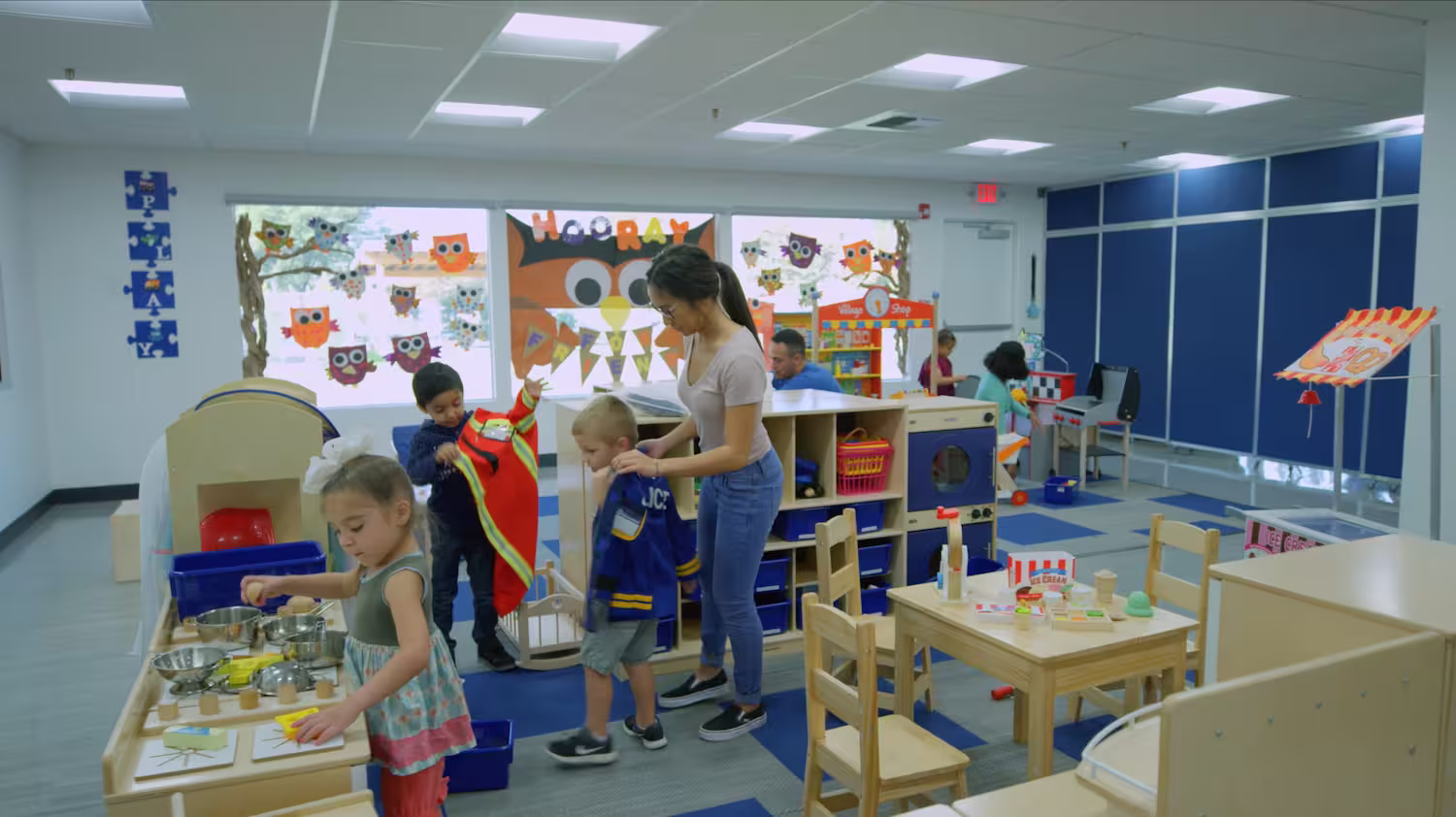 Children playing and a teacher helping a child dress up in a colorful classroom with owl decorations.