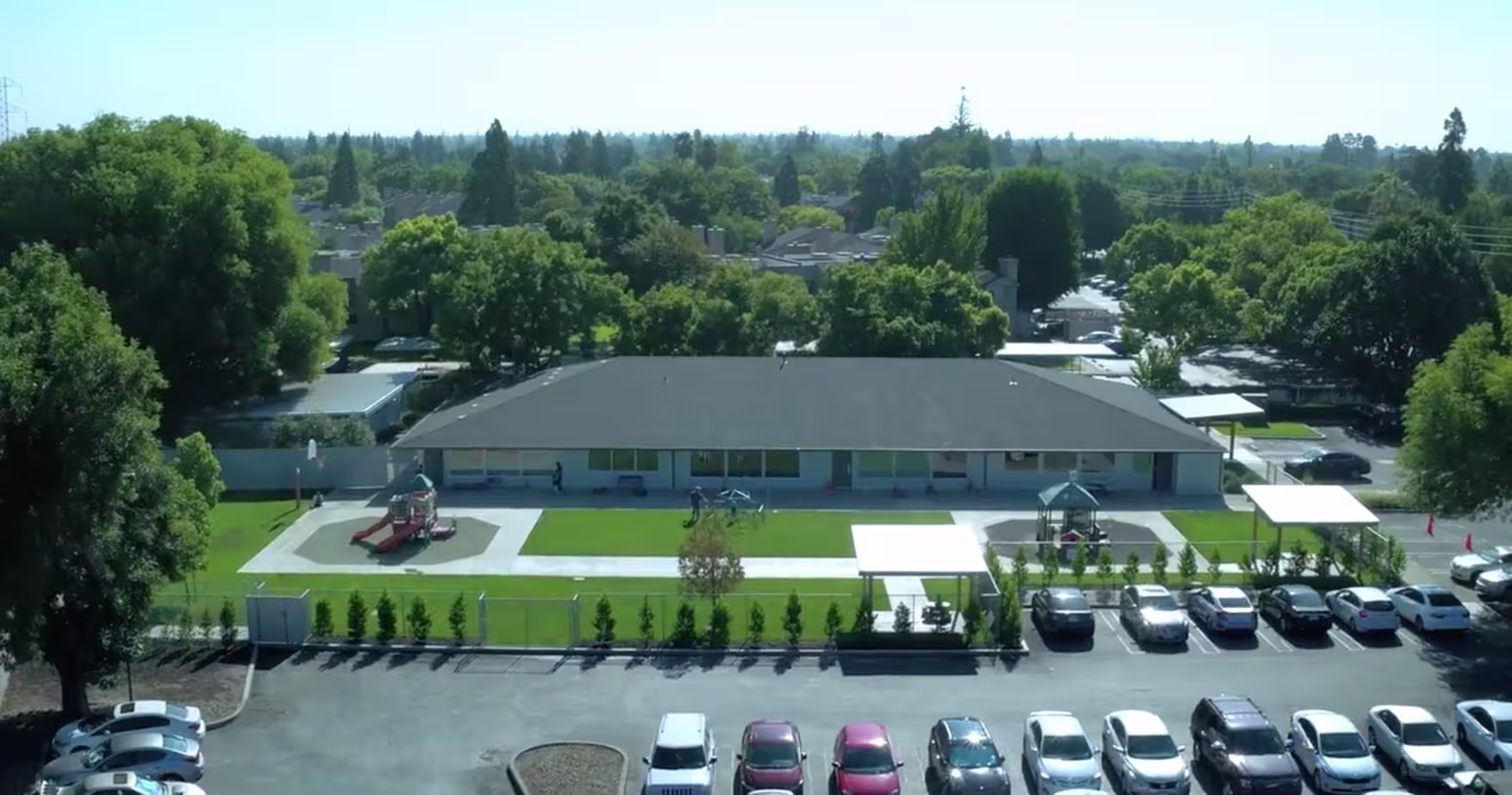 Aerial view of a building with a playground, green lawn, and parked cars in front and surrounding trees.