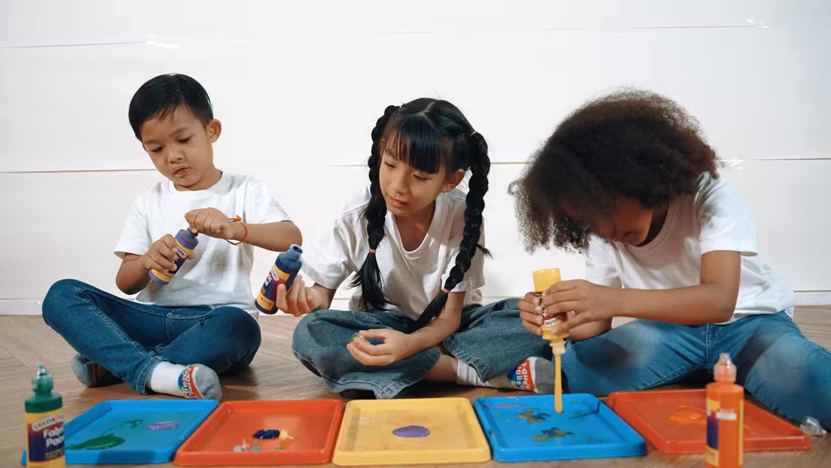 Three children sitting on floor painting on trays with colorful bottles of paint.